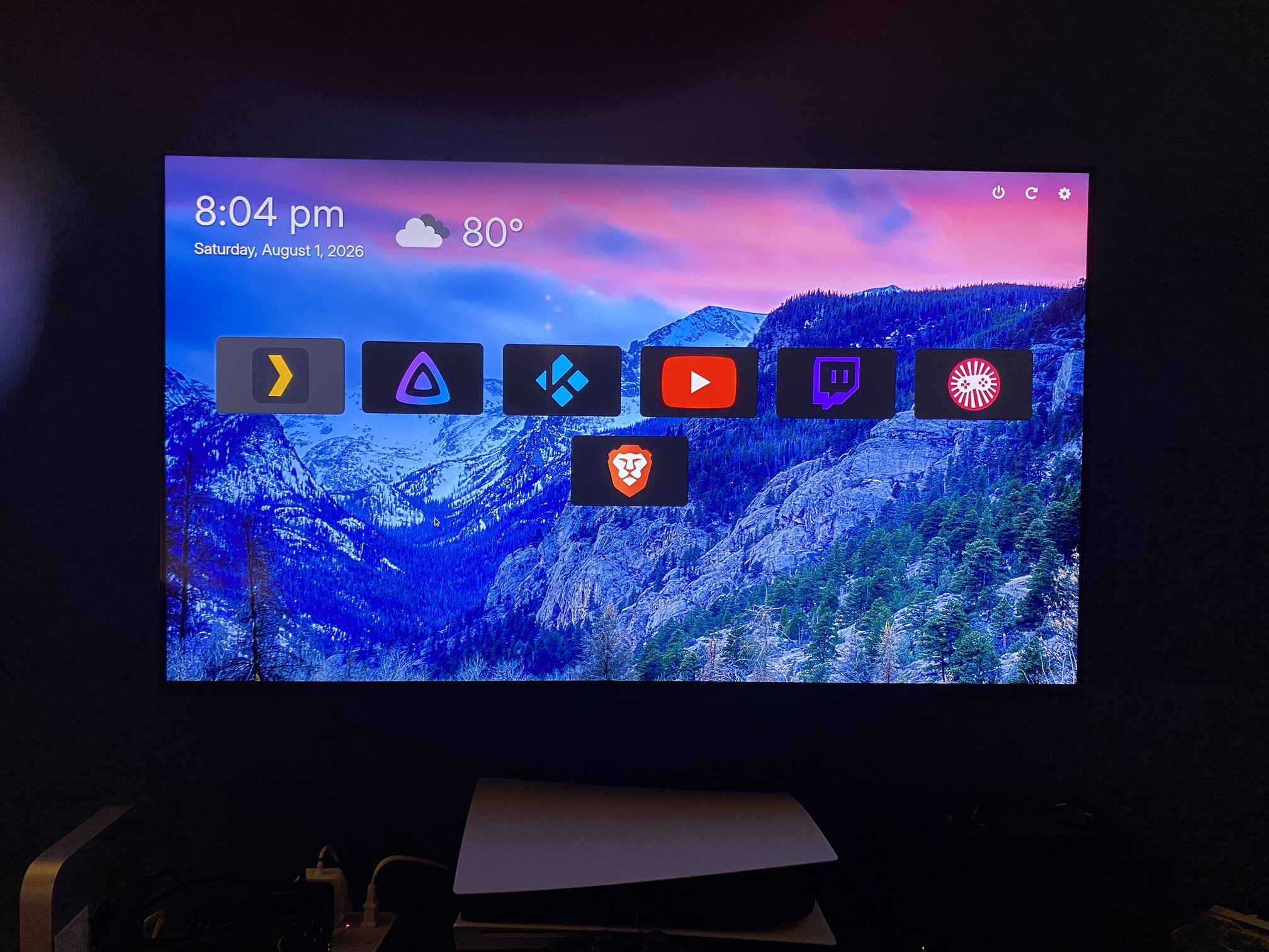 Photo of livingroom TV displaying the Smart Dashboard. A row of rectactular buttons and icons for various streaming services, apps and tools. Blueish background photo of a mountain.