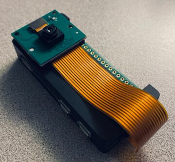 Raspberry Pi Zero W in a black metal case. Camera module is stuck onto the outside with double sided foam tape